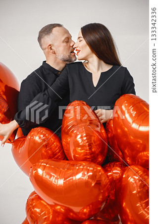 Romantic couple posing for a photo in anniversary. Studio full length shot of man embracing his wife and holding bunch of heart-shaped red ballons. Brunette woman and bearded man wearing black clothes 133342336