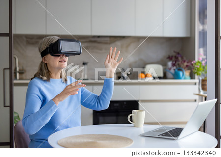 Senior woman wearing a vr headset and interacting with virtual space, sitting at a kitchen table with a laptop, embracing modern technology and the metaverse Senior woman wearing a vr headset and interacting with virtual space, sitting at a kitchen table with a laptop, embracing modern technology and the metaverse 133342340