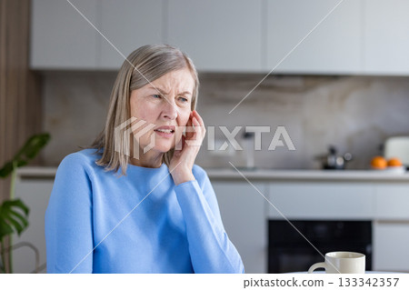 Senior woman suffering from a painful earache, touching her ear with a distressed expression while experiencing an uncomfortable health problem at home in her kitchen Senior woman suffering from a painful earache, touching her ear with a distressed expression while experiencing an uncomfortable health problem at home in her kitchen 133342357