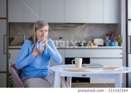 Senior woman feeling unwell and experiencing a painful cough with a hand on her chest while sitting at a kitchen table, indicating respiratory health issues or sickness Senior woman feeling unwell and experiencing a painful cough with a hand on her chest while sitting at a kitchen table, indicating respiratory health issues or sickness 133342364