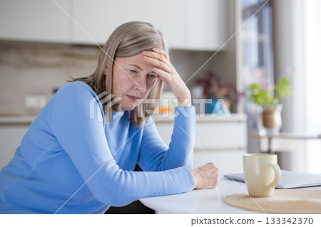Senior grey-haired woman seated at a kitchen table looking down with hand on forehead, appearing pensive and distressed, suffering from headache, worry and loneliness 133342370