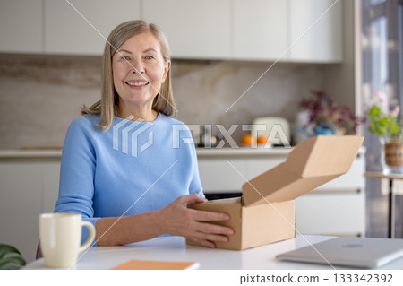 Senior woman smiling while unboxing a cardboard delivery package received from online shopping, sitting at a kitchen table with a laptop, representing e-commerce and consumer lifestyle Senior woman smiling while unboxing a cardboard delivery package received from online shopping, sitting at a kitchen table with a laptop, representing e-commerce and consumer lifestyle 133342392