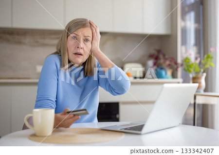 Senior woman at home holding a credit card and laptop, experiencing stress and confusion while attempting online shopping or banking, reacting to a potential fraud or scam 133342407
