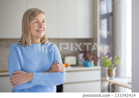 Senior woman looking away smiling, standing with arms crossed in her bright kitchen, experiencing happiness and positive emotions in her comfortable home Senior woman looking away smiling, standing with arms crossed in her bright kitchen, experiencing happiness and positive emotions in her comfortable home 133342414