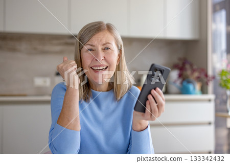 Senior woman is happily celebrating and expressing joy while looking at the camera, holding a smartphone and clenching her fist in excitement, reacting to good news, a win, or a successful outcome Senior woman is happily celebrating and expressing joy while looking at the camera, holding a smartphone and clenching her fist in excitement, reacting to good news, a win, or a successful outcome 133342432