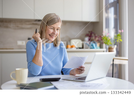 Senior woman smiling at kitchen table, reviewing bills and documents while using a laptop to budget, manage savings and plan a secure, confident retirement future 133342449