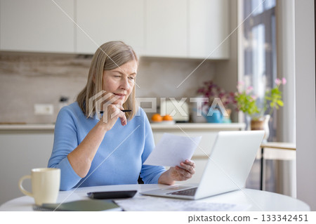 Mature woman analyzing financial documents and bills, calculating expenses at her kitchen table while using a laptop for budgeting and planning retirement savings 133342451
