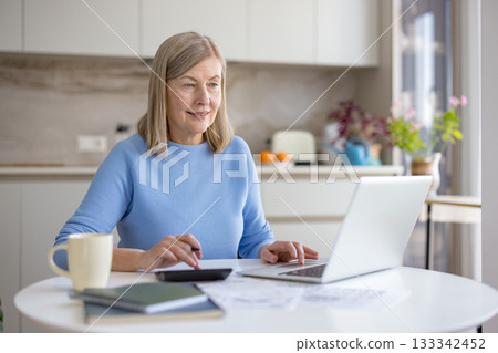 Mature woman at kitchen table managing household finances balancing bills, calculator and laptop, focused on budgeting, retirement planning and online accounting in a comfortable home setting 133342452