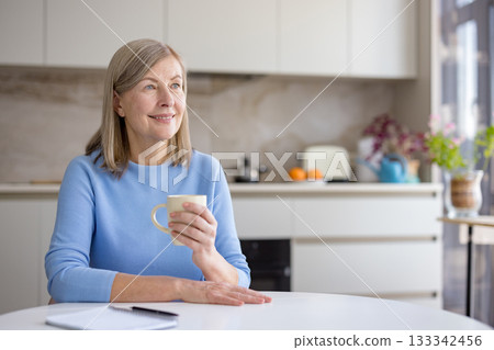 Senior woman relaxing at the kitchen table, holding a coffee mug, looking away with a thoughtful expression, enjoying a quiet moment at home during the morning 133342456