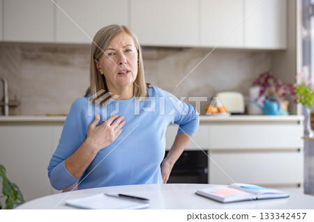 Senior woman at home experiencing a sudden chest pain and discomfort, holding her hand to her chest, possibly indicating a symptom of heart attack or heart disease 133342457