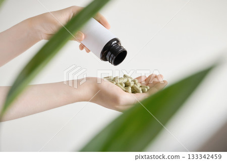 Female hands receiving herbal supplement capsules from bottle next to green plant 133342459