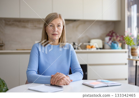 Mature businesswoman sitting at a table in her kitchen, engaging in a corporate video conference, representing remote work and freelance opportunities for older professionals Mature businesswoman sitting at a table in her kitchen, engaging in a corporate video conference, representing remote work and freelance opportunities for older professionals 133342465