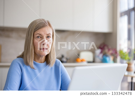 Senior woman focusing on her laptop screen, engaged in remote work or online activities while sitting in a bright, modern kitchen environment at her home Senior woman focusing on her laptop screen, engaged in remote work or online activities while sitting in a bright, modern kitchen environment at her home 133342478
