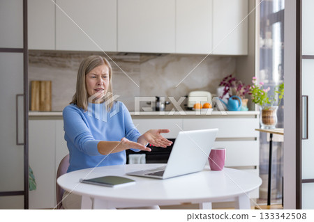 Senior woman feeling confused and overwhelmed while trying to use a laptop for a video call, gesturing in frustration with modern technology issues in her kitchen 133342508