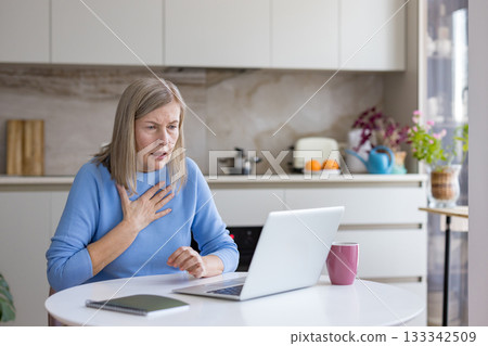 Senior woman suffering from sudden pain or anxiety, touching her chest and looking worriedly at the laptop screen during a virtual conversation in her kitchen Senior woman suffering from sudden pain or anxiety, touching her chest and looking worriedly at the laptop screen during a virtual conversation in her kitchen 133342509