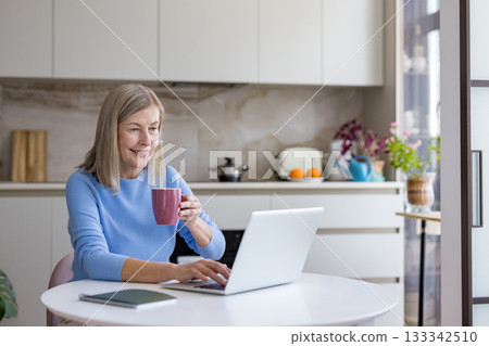 Senior woman enjoying remote work on a laptop at a kitchen table, holding a cup while typing, representing modern flexible employment and digital connectivity for mature adults Senior woman enjoying remote work on a laptop at a kitchen table, holding a cup while typing, representing modern flexible employment and digital connectivity for mature adults 133342510