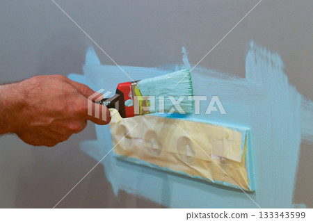 Using a brush to paint around an electrical socket on the wall, with masking tape applied for protection, as a DIY interior repair, aiming for precise results and safety. 133343599