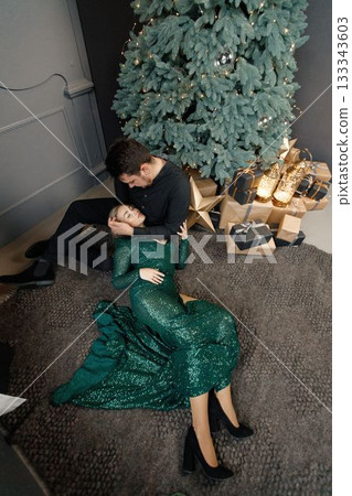 Young woman and man standing near christmas tree and hugging. Woman lay on her husband's lap. Blonde woman wearing fashion green dress and bearded man wearing black shirt. 133343603
