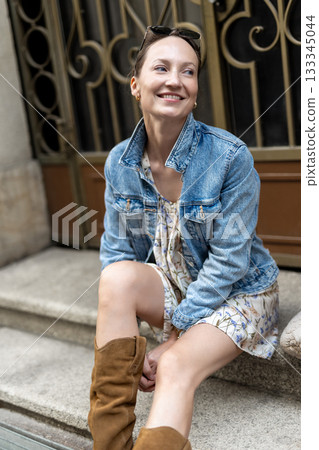 Portrait of young adult woman wear floral dress denim jacket brown suede boots sitting on stairs outdoors in old European city. Stylish female person enjoy walk travel urban lifestyle summertime day Portrait of young adult woman wear floral dress denim jacket brown suede boots sitting on stairs outdoors in old European city. Stylish female person enjoy walk travel urban lifestyle summertime day 133345044