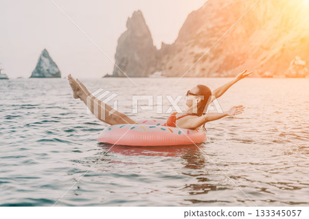Woman, inflatable, sea: happy woman floats on a donut ring, enjoying summer vacation freedom against scenic cliffs at sunset. Woman, inflatable, sea: happy woman floats on a donut ring, enjoying summer vacation freedom against scenic cliffs at sunset. 133345057