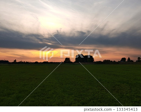 Dramatic Sunset Over Green Field with Silhouetted Trees. Stunning sunset with warm orange and pink sky over vast green field. Dark silhouettes of trees and dramatic clouds on horizon. 133345413