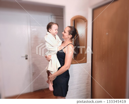 Happy Mother Holding Toddler After Bath. Young smiling mother in black dress tenderly holding her happy toddler wrapped in white towel after bath time. Warm family moment indoors. Happy Mother Holding Toddler After Bath. Young smiling mother in black dress tenderly holding her happy toddler wrapped in white towel after bath time. Warm family moment indoors. 133345419