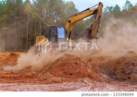 Excavator machinery digs into earth at construction site, sending dust into air among trees. 133346094