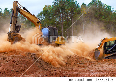 Construction skid steer equipment, excavator digs into ground, kicking up dust at site preparation land development. 133346106