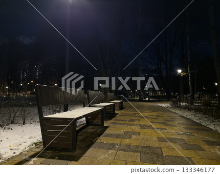 The first snow in the evening park. Snow covered wooden benches illuminated by streetlights. Park landscape with benches in snow and frozen trees. The first evening in the park after snow fell The first snow in the evening park. Snow covered wooden benches illuminated by streetlights. Park landscape with benches in snow and frozen trees. The first evening in the park after snow fell 133346177