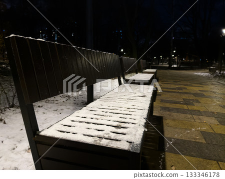 The seat of wooden bench is covered with the first snow of the year. Evening park with streetlights shining on the frozen bench, covered in ice and snow. It's impossible to sit on the cold bench The seat of wooden bench is covered with the first snow of the year. Evening park with streetlights shining on the frozen bench, covered in ice and snow. It's impossible to sit on the cold bench 133346178