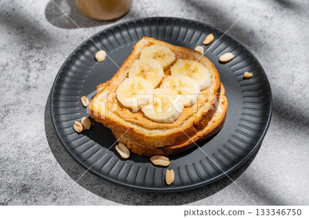 Peanut butter toast and banana on a black plate on a gray background with nuts 133346750
