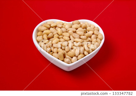 peeled pine nuts in a white heart-shaped bowl on a red background. peeled pine nuts in a white heart-shaped bowl on a red background. 133347782