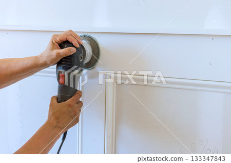 Carpenter carefully operating sander on white wood paneling during home improvement task. 133347843