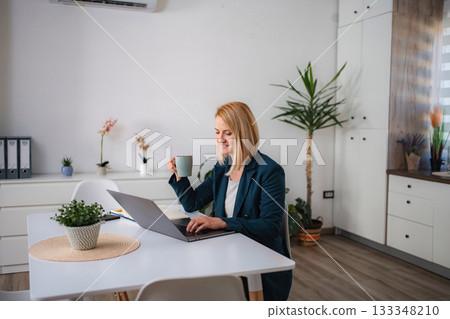 Smiling woman working from home using laptop and drinking coffee 133348210