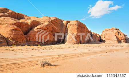 4wd vehicle tire tracks in sand desert, large rocky massif distance, clear blue sky above - typical scenery of Wadi Rum, Jordan 133348867