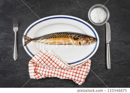 Whole smoked mackerel fish on white oval plate, gray black stone like table under. View from above Whole smoked mackerel fish on white oval plate, gray black stone like table under. View from above 133348875