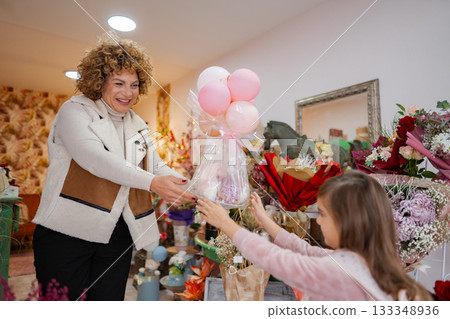 Curly-haired woman surprises a young girl with a colorful bouquet and balloons in a vibrant shop Curly-haired woman surprises a young girl with a colorful bouquet and balloons in a vibrant shop 133348936