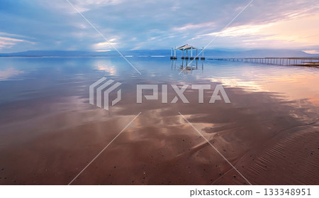 Calm dead sea surface in the morning, pink and blue sky above, wide angle photo, focus detail on sand foreground, blurred sun shelter background, typical scenery at Ein Bokek beach 133348951