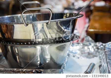 Shiny stainless steel mixing bowl and colander at a food stall, surrounded by foil, utensils, and pastry trays. Vendors prepare warm seasonal treats in a busy, atmosphere of festive market Shiny stainless steel mixing bowl and colander at a food stall, surrounded by foil, utensils, and pastry trays. Vendors prepare warm seasonal treats in a busy, atmosphere of festive market 133349013