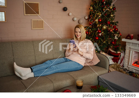 Cozy evening at home with a young woman relaxing by the Christmas tree while using her smartphone 133349669