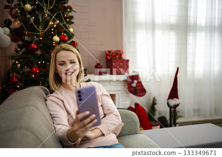 Joyful woman capturing holiday memories with her smartphone by a beautifully decorated Christmas tree 133349671