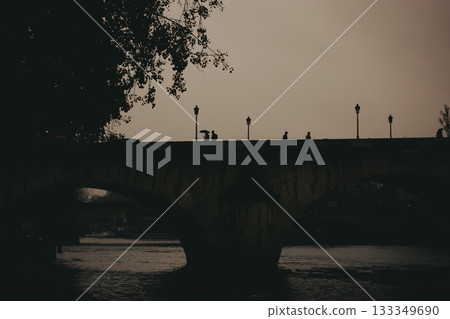 Silhouetted figures stroll across a bridge in Paris at dusk. The image captures a serene scene of people walking along a bridge in Paris, France, as the sun sets, creating a beautiful silhouette. Silhouetted figures stroll across a bridge in Paris at dusk. The image captures a serene scene of people walking along a bridge in Paris, France, as the sun sets, creating a beautiful silhouette. 133349690