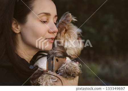 Woman lovingly embraces her small dog outdoors. A tender moment of affection. A girl is holding Yorkshire terrier doggy, enjoying a peaceful moment. Woman lovingly embraces her small dog outdoors. A tender moment of affection. A girl is holding Yorkshire terrier doggy, enjoying a peaceful moment. 133349697