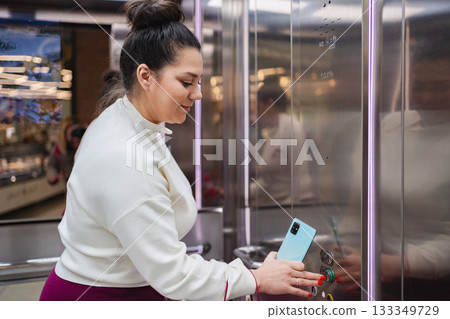 Woman pressing elevator button while using smartphone in a mall Woman pressing elevator button while using smartphone in a mall 133349729