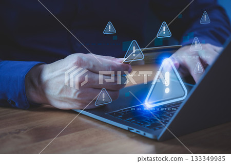Person holding a credit card while using a laptop computer with warning symbol, representing online payment risk, online shopping, financial fraud, and identity theft during digital transactions. 133349985