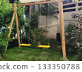 Wooden swing set with two yellow seats hangs from chains, surrounded by lush greenery and plants. The area features a grass-covered floor, providing a vibrant, natural ambiance 133350788