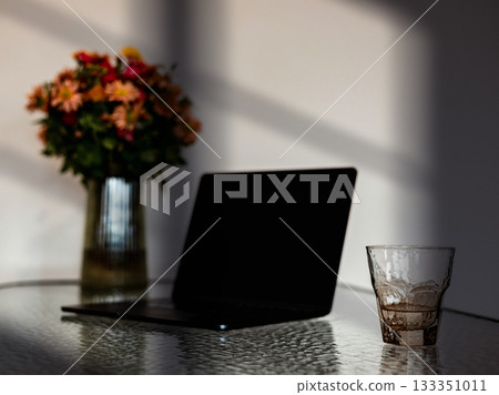 Cozy home workspace with laptop, glass of water, and a vase of flowers in warm natural light. Remote-work in calm homy atmosphere. Morning light. 133351011