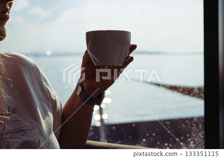 A woman enjoying a cup of coffee by the water. A person holds a white mug near a window with calm sea view, a peaceful serene atmosphere. Morning in cafe, restaurant terrace balcony. Relaxing people. A woman enjoying a cup of coffee by the water. A person holds a white mug near a window with calm sea view, a peaceful serene atmosphere. Morning in cafe, restaurant terrace balcony. Relaxing people. 133351115