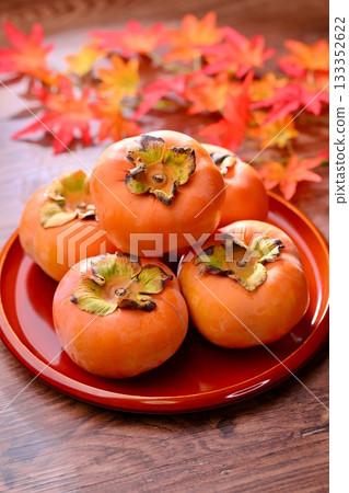 Persimmon, a Japanese autumn fruit. The maple leaves in the background are artificial. Persimmon, a Japanese autumn fruit. The maple leaves in the background are artificial. 133352622