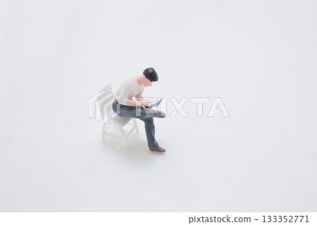 Miniature Man Reading a Book While Seated on a White Chair Miniature Man Reading a Book While Seated on a White Chair 133352771